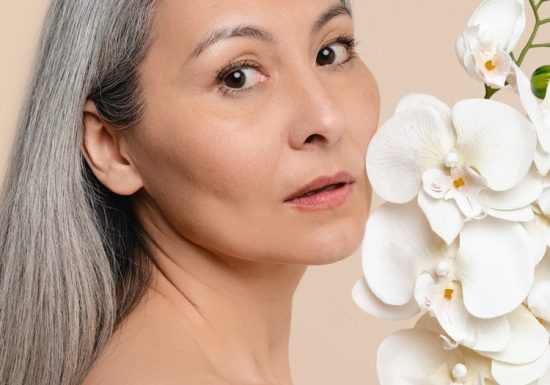 Beautiful mature middle-aged woman shirtless naked looking at camera, holding orchid flower isolated in beige background. Rejuvenation concept. Aging skin hair care. Cosmetics